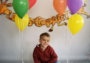 Chłopiec siedzi na tle fotograficznym i pozuje do zdjęcia wśród balonów.